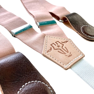May include: A pair of light pink suspenders with brown leather accents. The suspenders have a metal clip on each end and a leather patch with a star and a logo in the center.