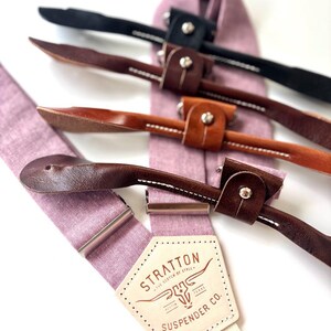 May include: Close-up of several suspenders with leather accents and metal hardware. The suspenders are in various colors, including black and brown, and are attached to a light purple fabric. The label reads "STRATTON SUSPENDER CO."