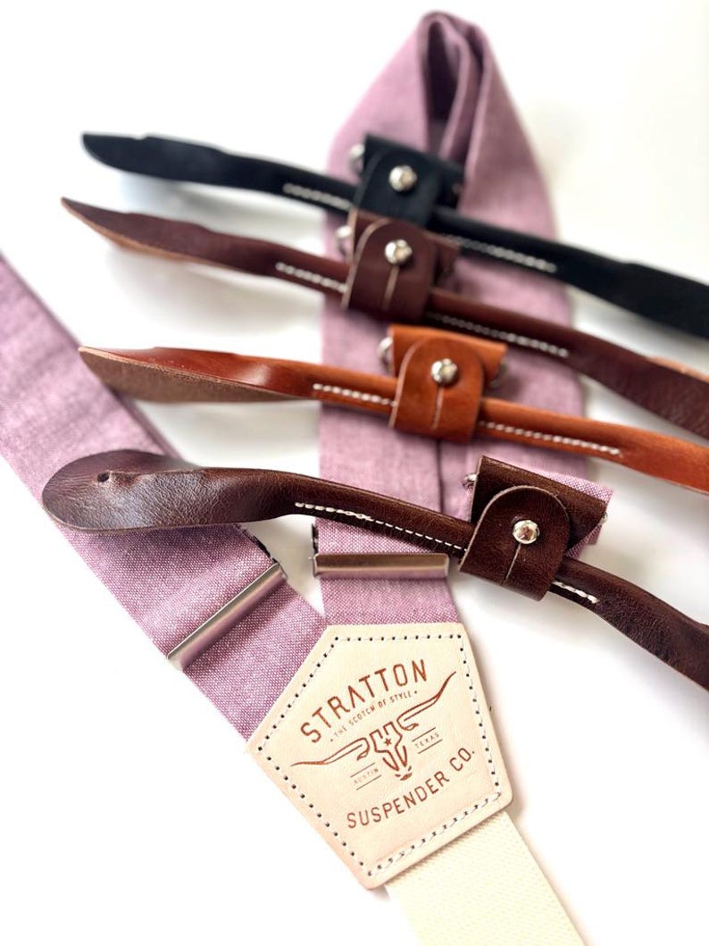 May include: Close-up of several suspenders with leather accents and metal hardware. The suspenders are in various colors, including black and brown, and are attached to a light purple fabric. The label reads "STRATTON SUSPENDER CO."