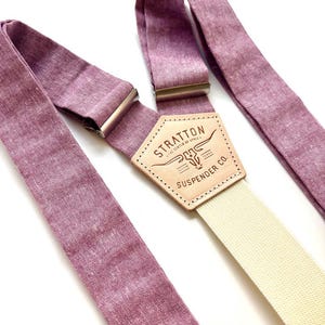 May include: A pair of mauve suspenders with a cream-colored strap and silver-tone metal hardware. The leather patch in the center features the text "STRATTON SUSPENDER CO." and a steer head logo.