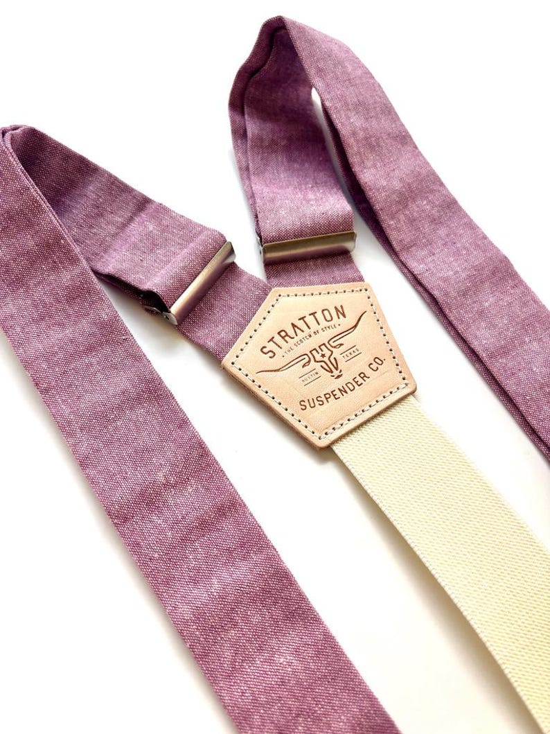 May include: A pair of mauve suspenders with a cream-colored strap and silver-tone metal hardware. The leather patch in the center features the text "STRATTON SUSPENDER CO." and a steer head logo.