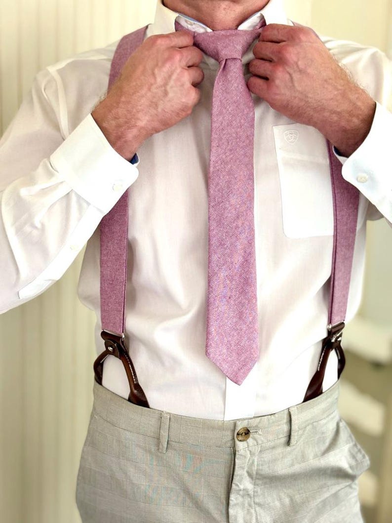 May include: A person adjusting a light purple tie, with matching suspenders. The tie and suspenders are made of a textured fabric. The person is wearing a white dress shirt and light gray pants.