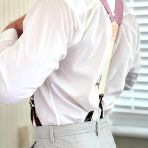 May include: A person wearing a white dress shirt and light gray trousers with suspenders. The suspenders are a light pink color with cream-colored straps and brown leather attachments. The suspenders have a small leather tag with text.