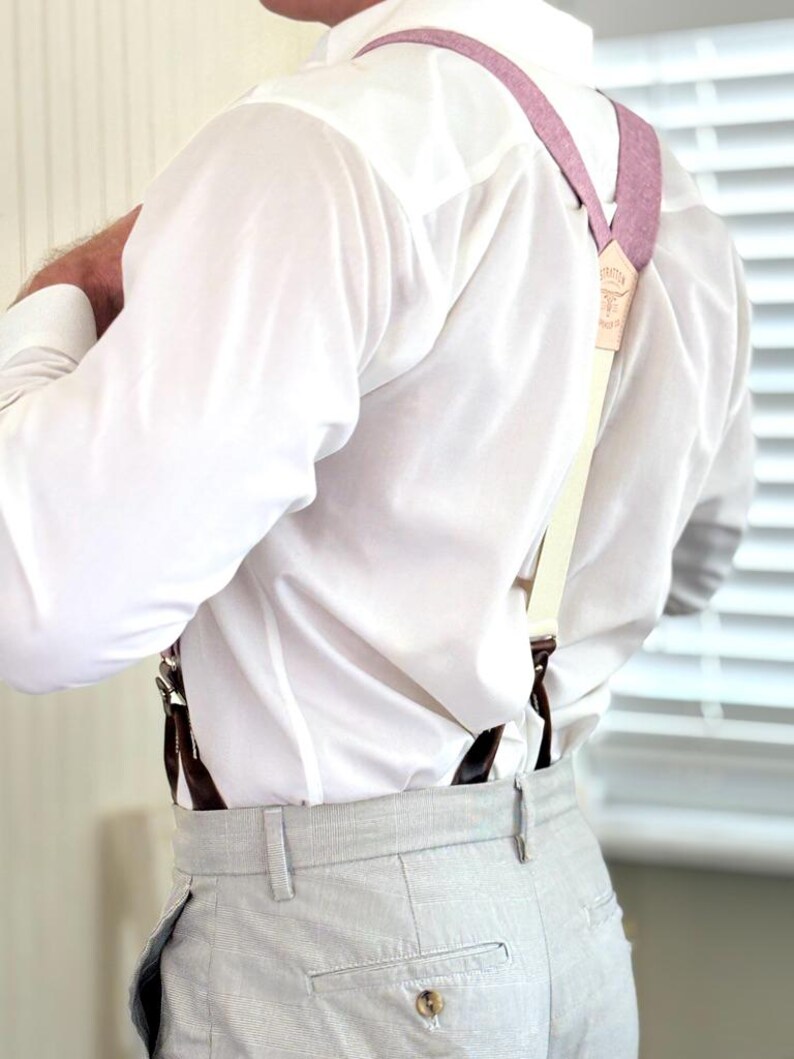 May include: A person wearing a white dress shirt and light gray trousers with suspenders. The suspenders are a light pink color with cream-colored straps and brown leather attachments. The suspenders have a small leather tag with text.
