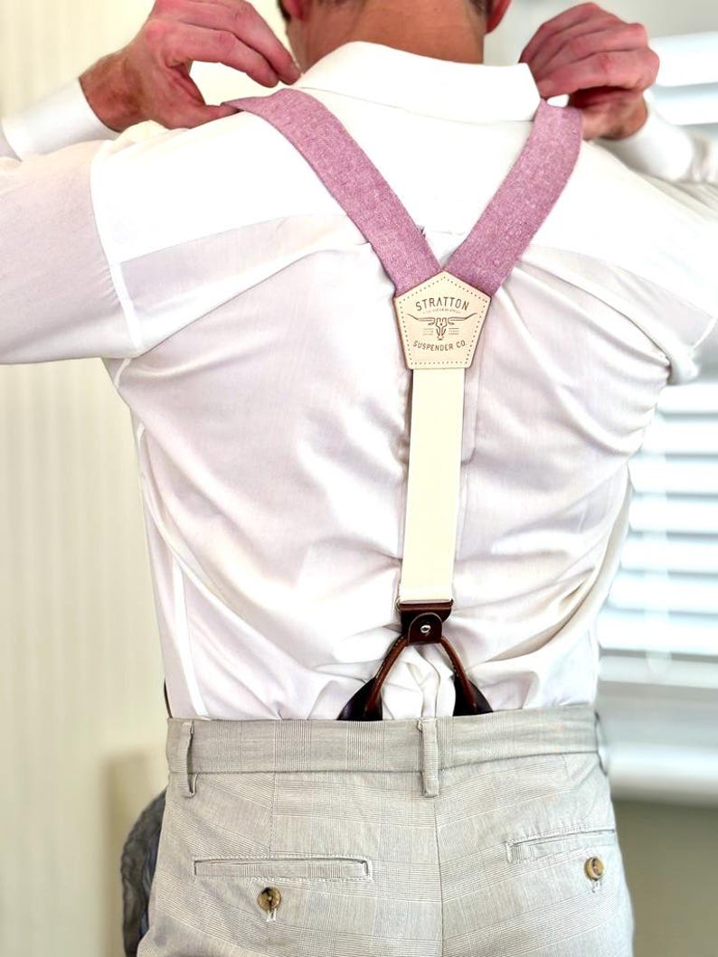May include: A person adjusting light pink suspenders with cream-colored straps and brown leather accents. The suspenders are attached to light gray plaid trousers. The suspenders have a leather patch with the text "STRATTON SUSPENDER CO."