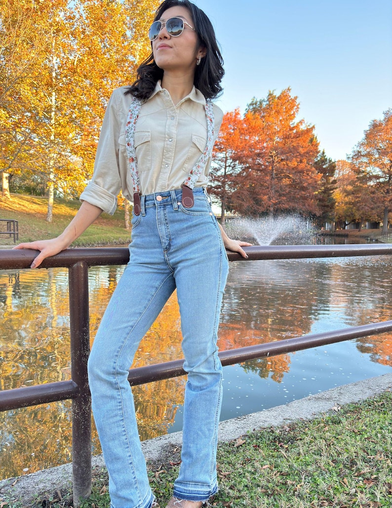 Women’s floral magnetic clasp suspenders with brown leather tabs, styled with blue jeans and a cream button-up shirt by a pond with fall trees.