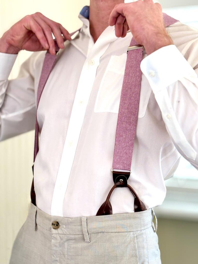 May include: A close-up of a person wearing a white dress shirt and light gray pants with pink suspenders. The suspenders have brown leather attachments and silver metal clasps. The shirt has a blue collar.