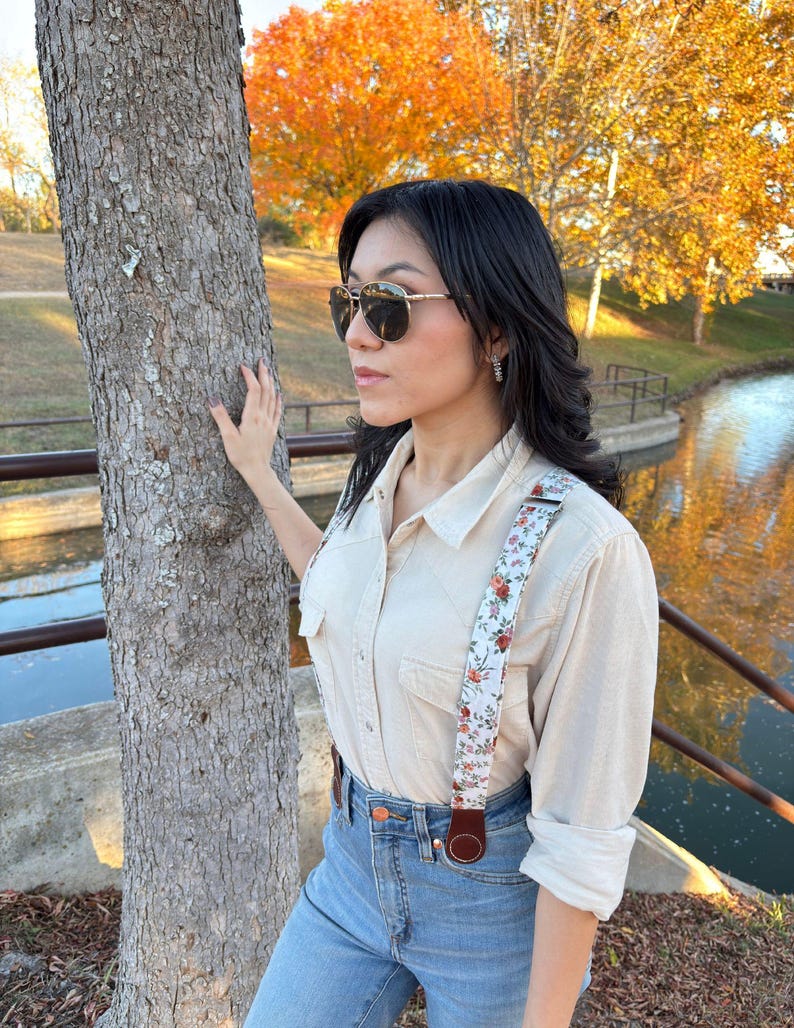 Close lifestyle view of floral women’s magnetic clasp suspenders with brown leather, worn with blue jeans & a cream shirt in autumn outdoor setting.
