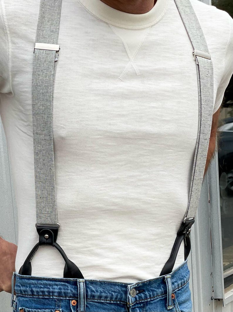 Men’s heather gray linen button-on suspenders paired with denim jeans and a classic off-white crewneck shirt. A perfect wedding and groomsmen accessory, these handcrafted suspenders blend vintage style with rugged craftsmanship. Ethically made in Texas by Stratton Suspender Co.