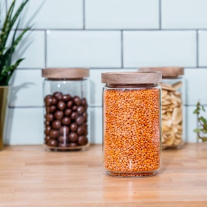 Peut inclure: Trois bocaux en verre avec des couvercles en bois, remplis d'articles différents. Le bocal de gauche contient des bonbons au chocolat, le bocal du milieu contient des lentilles orange et le bocal de droite contient des fruits secs.