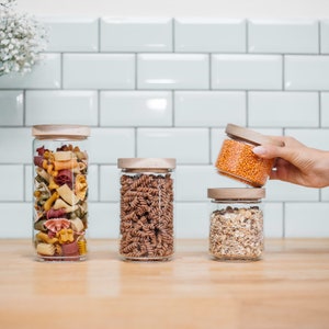 May include: Four glass jars with wooden lids filled with different types of pasta and grains. The jars are on a wooden worktop. One jar is being held by a hand.