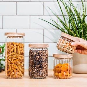 Peut inclure: Quatre bocaux en verre avec des couvercles en bois, remplis de différents produits de base. Les bocaux ont un design côtelé et sont remplis de pâtes, de fruits secs, de graines et d'herbes séchées.