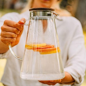 Könnte beinhalten: Eine klare Glaskaraffe mit einem Holzdeckel und einem Edelstahlsieb. Die Karaffe ist mit Wasser und Orangenscheiben gefüllt.