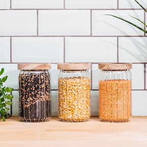 May include: Three glass jars with wooden lids filled with different types of dried beans. The jars are sitting on a wooden surface in front of a white tiled wall.