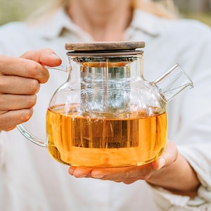 Könnte beinhalten: Eine klare Glas-Teekanne mit Holzdeckel und Edelstahl-Infuser. Die Teekanne ist mit bernsteinfarbenem Tee gefüllt. Die Teekanne hat einen gebogenen Griff und einen Ausguss. Das Bild zeigt ein Teeservice.