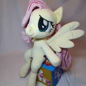 May include: A plush toy of a yellow pony with a light pink mane and tail, featuring large embroidered eyes and wings. The pony is sitting on a small, square, blue and yellow block decorated with pixelated character graphics on its sides.