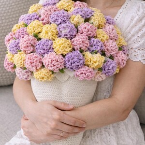 May include: A handmade crochet bouquet in a cream-coloured cone-shaped vase. The bouquet features a variety of colourful crochet flowers in shades of pink, yellow, and purple. The vase is held by a person.