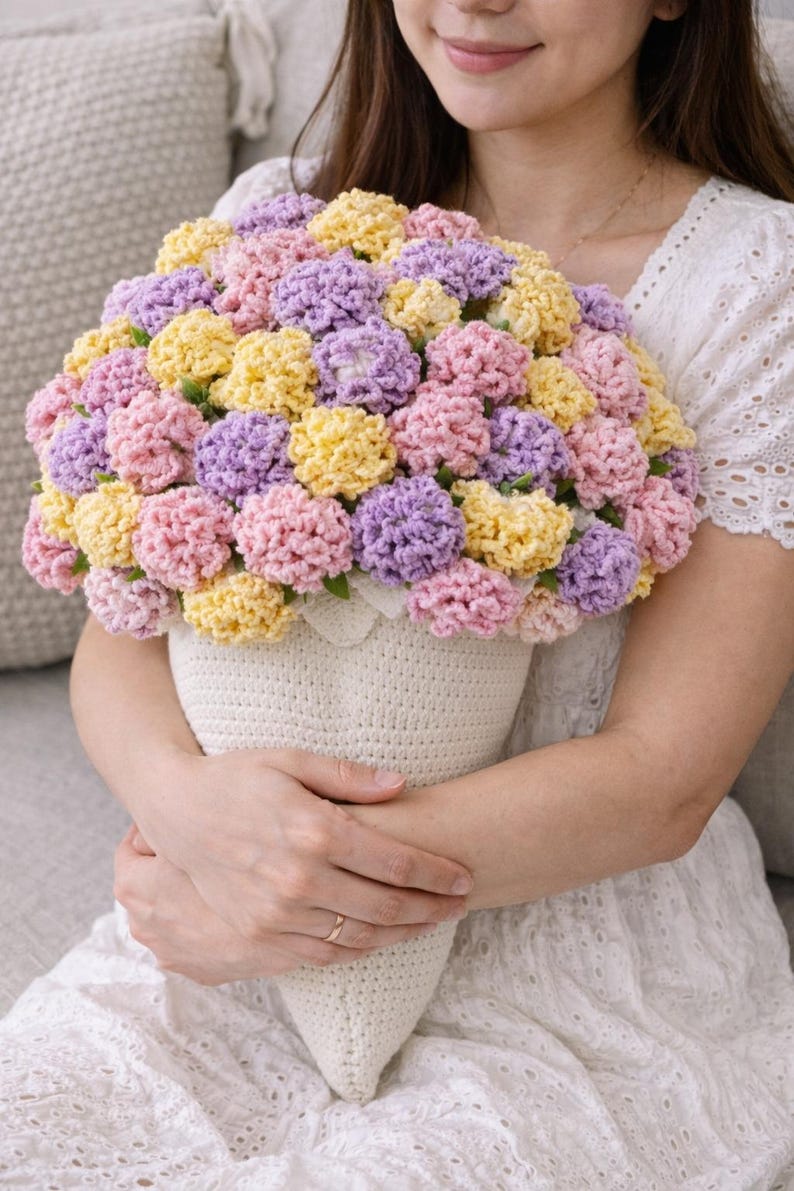 May include: A handmade crochet bouquet in a cream-coloured cone-shaped vase. The bouquet features a variety of colourful crochet flowers in shades of pink, yellow, and purple. The vase is held by a person.