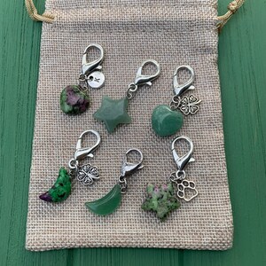 May include: A set of six green gemstone charms with silver clasps and charms. The charms include a heart, a star, a crescent moon, a four-leaf clover, a paw print, and a star. The charms are displayed in a small burlap bag.