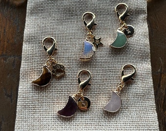 May include: Five gold charm pendants with crescent moon shapes and different colored stones. The charms have a gold lobster clasp and a small decorative charm attached to the top of the crescent moon. The charms are displayed on a beige fabric surface.