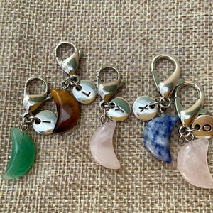 May include: Four silver keychains with crescent moon charms. Each charm is made of a different stone: green aventurine, rose quartz, blue sodalite, and tiger's eye. Each keychain also has a silver charm with a letter on it: L, Y, X, and O.