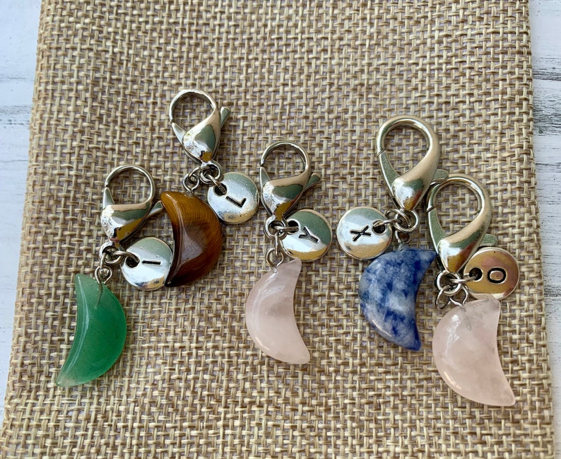 May include: Four silver keychains with crescent moon charms. Each charm is made of a different stone: green aventurine, rose quartz, blue sodalite, and tiger's eye. Each keychain also has a silver charm with a letter on it: L, Y, X, and O.