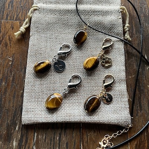 Puede incluir: Cinco dijes de ojo de tigre color dorado con cierres plateados y un collar de cordón negro. Los dijes están sobre un fondo de tela beige.