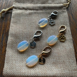 Op de afbeelding: Drie ovale opaalcharms met zilveren en gouden sluitingen en bedels. De charms liggen op een jute achtergrond.