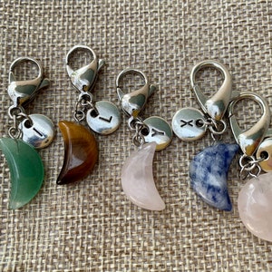 May include: Five silver keychains with crescent moon charms made of different stones. The stones are green, brown, pink, blue, and rose quartz. Each keychain has a silver charm with a letter on it.