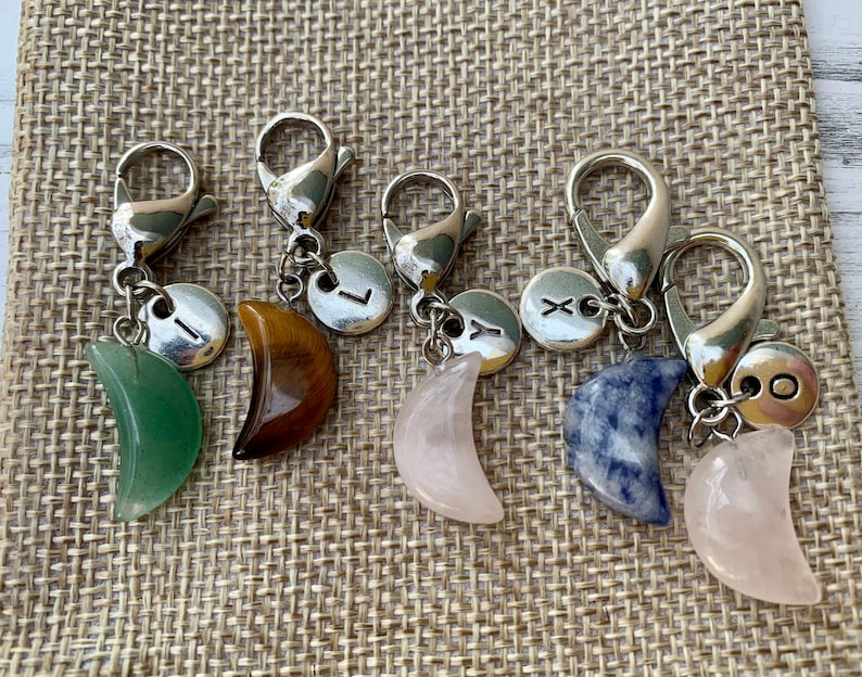 May include: Five silver keychains with crescent moon charms made of different stones. The stones are green, brown, pink, blue, and rose quartz. Each keychain has a silver charm with a letter on it.