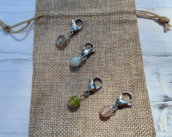 May include: Four silver and crystal bead stitch markers with silver lobster clasps. The beads are clear, white, green, and pink.