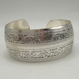 May include: A wide silver-toned cuff bracelet featuring an open back design. The surface is decorated with two horizontal bands of intricate, raised scrollwork patterns separated by a smooth central strip and bordered by fine beaded detailing.