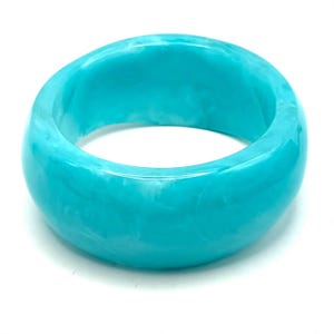 May include: A thick, rounded bangle bracelet in a vibrant turquoise blue color. The material features a smooth, polished finish with subtle, swirling white marbled patterns throughout the surface, creating a continuous, solid circular form.