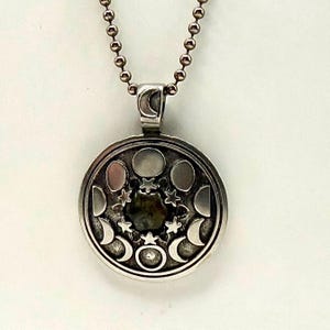 May include: A silver-toned circular pendant necklace featuring a central dark stone surrounded by small star shapes and a series of moon phases engraved around the perimeter. The pendant hangs from a matching silver-toned ball chain necklace.