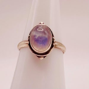 May include: A silver ring featuring an oval, iridescent gemstone with shades of purple and blue. The gemstone is set in a decorative bezel, and the ring has a simple, polished band. The ring is displayed on a white surface.