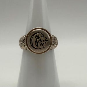 May include: A silver-toned ring featuring a circular face with a dark, etched crescent moon and floral design. The band displays an intricate, textured pattern along its sides, providing a detailed appearance to the metallic jewelry piece.