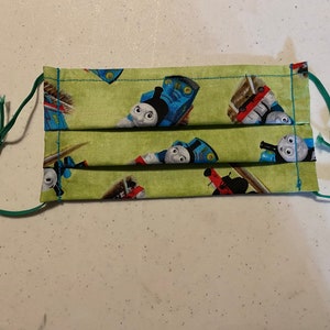 May include: A green face mask with a repeating pattern of Thomas the Tank Engine characters. The mask has elastic ear loops.