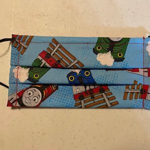 May include: A blue fabric face mask with a repeating pattern of cartoon trains on a white background. The mask has black elastic ear loops.