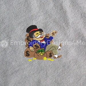 Puede incluir: Un diseño bordado de un hombre Monopoly de dibujos animados sentado en una pila de bolsas de dinero. Lleva un sombrero de copa, un monóculo y una corbata de lazo. Las bolsas de dinero son marrones con signos de dólar y dinero verde en su interior. Hay monedas de oro esparcidas alrededor de las bolsas.