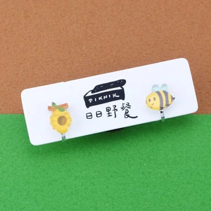 May include: Clip-on earrings featuring a yellow sunflower and a cartoon bee with black and yellow stripes. The earrings are displayed on a white card with the word "PIKNIK" and Japanese text. The background is brown and green.
