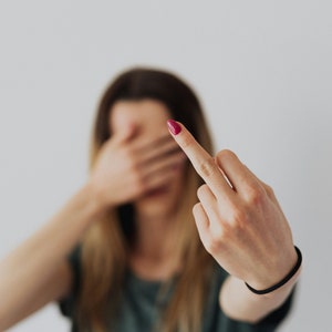 May include: A person with long brown hair is out of focus in the background. The person's hand is in focus and is giving the middle finger. The person's fingernail is painted a dark pink color.