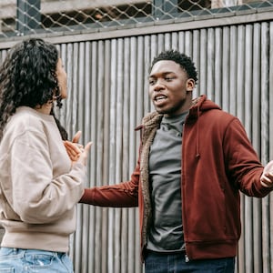 Op de afbeelding: Een man en een vrouw voeren een gesprek. De man draagt een rode hoodie over een grijs t-shirt en een blauwe jeans. De vrouw draagt een beige trui en een blauwe jeans. Ze staan voor een houten muur.