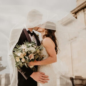 May include: A couple kissing under a sheer white veil. The person on the left is wearing a black tuxedo with a bow tie, and the person on the right is wearing a white wedding dress and holding a bouquet of flowers.