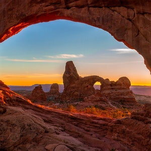May include: A view of a natural sandstone arch in Arches National Park, Utah. The arch is framed by a larger rock formation, and the view through the arch shows a distant mountain range at sunset.