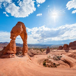 May include: A large, red sandstone arch stands tall against a bright blue sky with fluffy white clouds. The arch is a natural formation in a desert landscape.