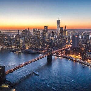 May include: An aerial view of the New York City skyline at dusk, with the Brooklyn Bridge in the foreground and the Freedom Tower in the background. The city lights are reflected in the water.