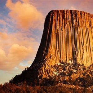 May include: A large, towering rock formation with a textured, striated surface. The rock is a light brown color and is set against a backdrop of a cloudy sky with shades of pink and orange.