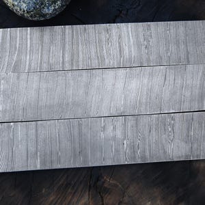 May include: Three rectangular metal billets featuring a distinct grey and silver layered pattern. Each piece displays a consistent, flowing grain texture across its flat surface, characteristic of Damascus steel construction.