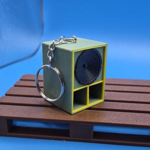 Puede incluir: Un llavero en forma de cubo que representa una caja de altavoz en miniatura de color verde oliva con un marco amarillo. La parte frontal muestra un cono de altavoz negro circular sobre dos puertos cuadrados. Un anillo de metal plateado está unido a la parte superior.