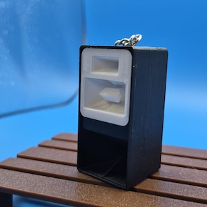 Op de afbeelding: Een zwarte rechthoekige sleutelhanger in de vorm van een luidsprekerkast met een wit voorpaneel met twee afzonderlijke openingen voor hoornluidsprekers. Het object bevat een kleine zilverkleurige metalen ring aan de bovenkant om op te hangen.